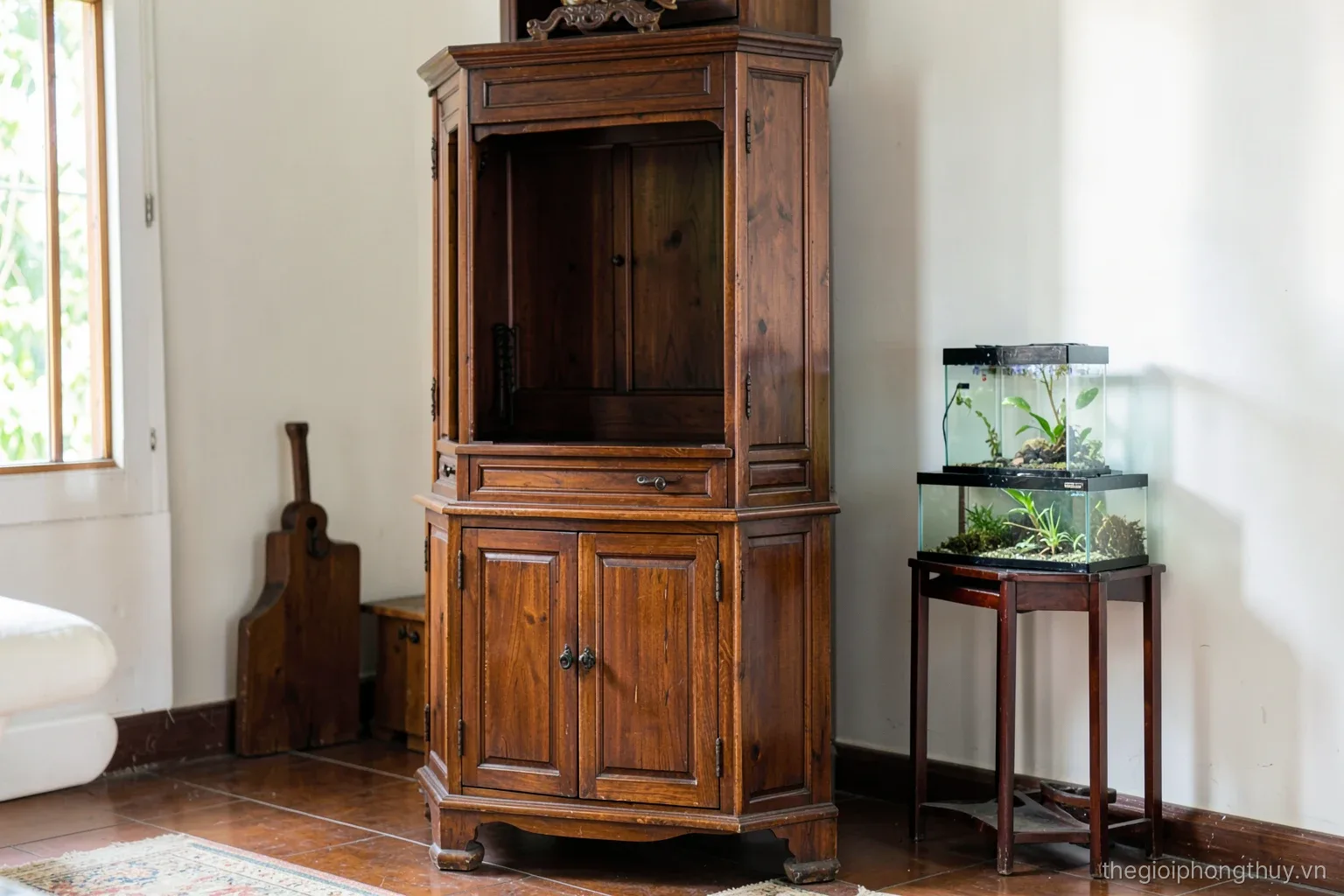 Side view of a tall wooden ancestral altar cabinet in a Vietnamese living room, closed lower cabinet doors, a small aquarium placed on a separate stand nearby, daylight